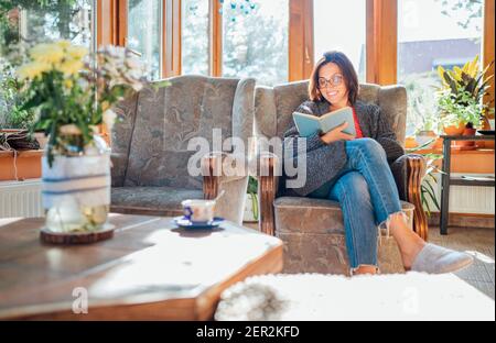 Allegra donna sorridente vestito accogliente abiti a casa seduta in comoda poltrona, rilassante, leggere un libro e bere caffè o tè del mattino in cous Foto Stock