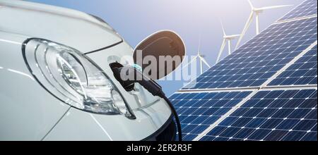Close up di auto elettrica collegata a un cavo di ricarica sullo sfondo dei pannelli solari e delle turbine a vento - fonti di energia pulita e rinnovabile Foto Stock