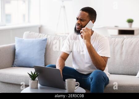 Uomo africano dispiaciuto che parla al telefono seduto al computer portatile indoor Foto Stock