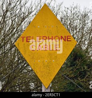 Segnale del cavo telefonico vecchio e intemperie. Giallo con testo rosso a forma di rombo. Alberi sfocati sullo sfondo. Foto Stock