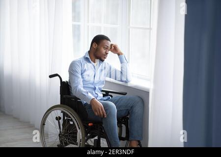 Insoddisfatto ragazzo nero handicappato in sedia a rotelle guardando fuori finestra, sensazione triste e disperato a casa, spazio copia Foto Stock