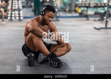 Sportivo nero rilassato che ascolta la musica sullo smartphone, interno della palestra Foto Stock