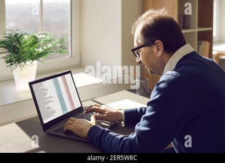 Uomo seduto alla scrivania dell'ufficio e che lavora con documenti aziendali e fogli di calcolo su computer portatile Foto Stock