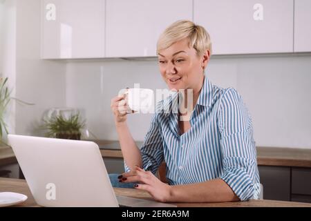 Giovane donna allegra che beve caffè e parla in videoconferenza sul computer portatile mentre si è seduti in cucina a casa Foto Stock