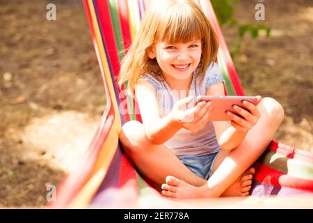 Sorridente bambino ragazza registrazione stile di vita blog parlare con la fotocamera del telefono cellulare all'aperto, seduta su amaca. Felice giovane influencer filmare vlog per il suo canale. Concetto di blogging. Foto Stock