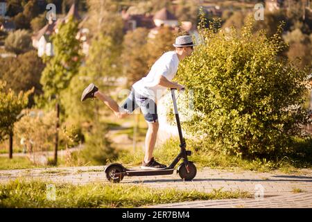 Uomo gioioso e casual in cappello e occhiali da sole che cavalcano elettrici scooter nel parco estivo della città Foto Stock