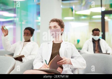Ritratto di giovane donna che indossa maschera e camice da laboratorio mentre si ascolta la lezione di medicina in università o centro di lavoro, copia spazio Foto Stock