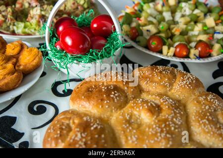 Tavolo pasquale con uova rosse colorate, pane, rami verdi decorati, tavolo in legno su tavola bianca con tovaglia tessile Foto Stock