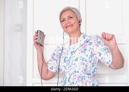 Una donna anziana dai capelli grigi in un abito vintage, in cuffie, con un radio-cassetta stereo portatile in mano che ascolta musica e balla Foto Stock