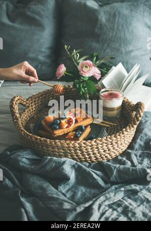 Prima colazione in camera. Cialde con arancia e mirtilli, latte di rosa in vetro e lacca con frizzante al miele nel vassoio del cesto sopra il letto di lino grigio scuro Foto Stock