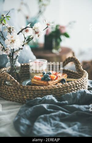 Prima colazione in camera. Waffle con arancio e mirtilli di sangue, latte di rosa in vetro e fiori bianchi in fiore nel vassoio del cesto sopra il letto di lino grigio scuro lin Foto Stock