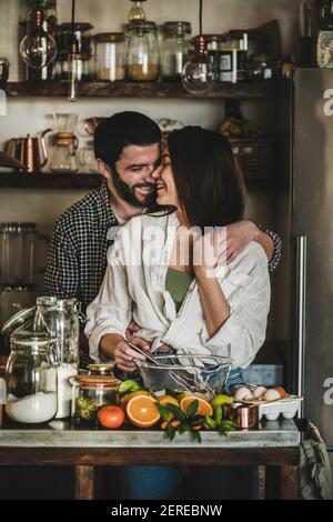 Giovane dolce famiglia coppia cucinare insieme in cucina. Giovane uomo caucasico e bruna donna abbracciando, flirtare, ridendo e facendo l'impasto per la cottura Foto Stock