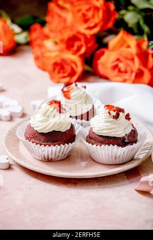 Cupcakes di velluto rosso fatto in casa con panna montata su piastra di ceramica rosa, tovagliolo bianco con nastro, fiori di rose, cuori di legno su fondo di tessitura rosa Foto Stock