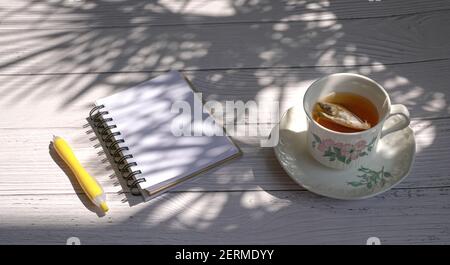 Tazza di tè con blocco note e penna gialla su tavolo di legno con ombra di foglie di palma. Foto Stock