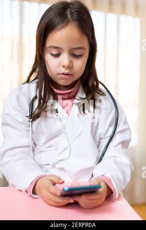 Ritratto di bambina vestita in uniforme da medico con uno smartphone. Concetto di tecnologia e benessere. Foto Stock