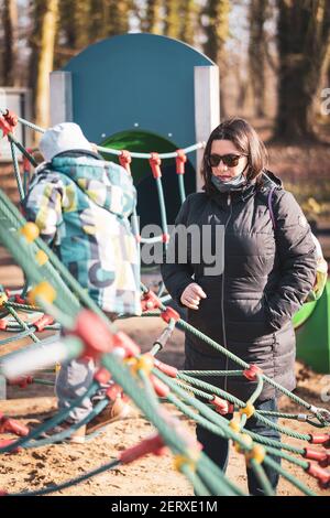 POZNAN, POLONIA - 21 feb 2021: Donna con occhiali da sole in piedi da un ragazzo su una griglia di corda attrezzatura per salire in un parco giochi nel Solacki Park. Relat Foto Stock