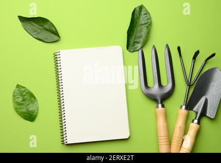 taccuino aperto con fogli bianchi vuoti e vari attrezzi da giardinaggio con maniglie in legno su sfondo verde, disposizione piatta, spazio di copia Foto Stock