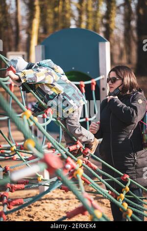 POZNAN, POLONIA - 21 feb 2021: Donna con occhiali da sole in piedi da un ragazzo su una griglia di corda attrezzatura per salire in un parco giochi nel Solacki Park. Relat Foto Stock