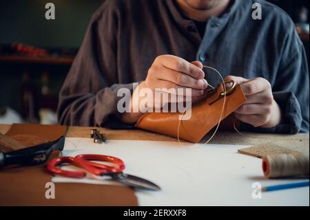 Seduto ad un tavolo in officina un ragazzo giovane cucita manualmente gli elementi in pelle l'uno sull'altro Foto Stock