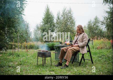 La ragazza si annoia e passa il tempo in social network durante un picnic. Preparazione della carne arrosto sulla griglia. C'è un tavolo con spuntini e birra Foto Stock