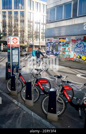 Le biciclette a noleggio Santander attendono i loro prossimi piloti, di fronte a un pendolarismo ciclistico a Shoreditch dove i graffiti coprono l'esterno di un ex ufficio, il 26 febbraio 2021, a Londra, Inghilterra. Foto Stock