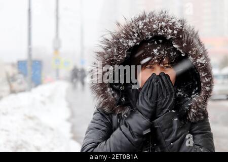 Tempo freddo e bizzard in città, donna in pelliccia cappuccio in piedi sulla strada invernale durante la nevicata Foto Stock