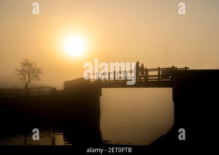 Incredibile luce arancione nebbia alba in coppia amore silhouette look vista in nebbia mindfulness tranquillo ponte di legno piedi sopra tranquillo canale fluviale Foto Stock