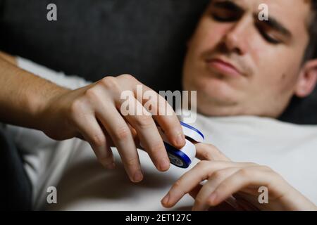 Un giovane in una T-shirt bianca è malato con il coronavirus COVID-19 e misura la saturazione di ossigeno a casa sul divano. Pulsossimetro portatile digitale Foto Stock