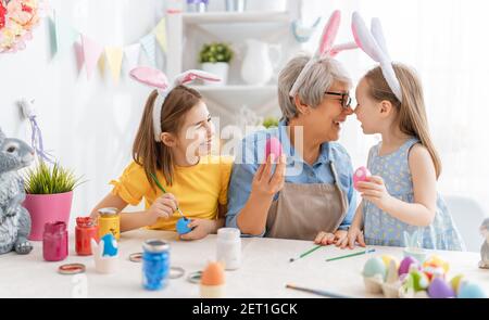 Una nonna e le sue nipotine dipingendo le uova. Buona famiglia che si prepara per Pasqua. Carino bambine che indossano orecchie conigliate. Foto Stock