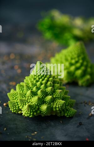 primo piano di alcune teste crude di broccoli di romanesco del bambino su una superficie scura della pietra spruzzata con erbe, spezie e semi differenti Foto Stock