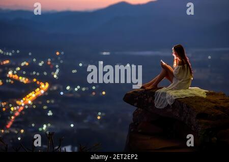Donna seduta su una sporgenza sopra la città utilizzando un telefono cellulare, Thailandia Foto Stock