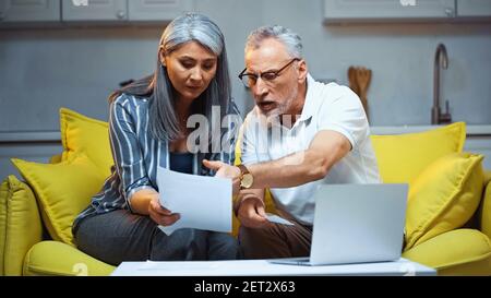 uomo serio che punta con la mano al documento vicino al portatile e. moglie asiatica Foto Stock