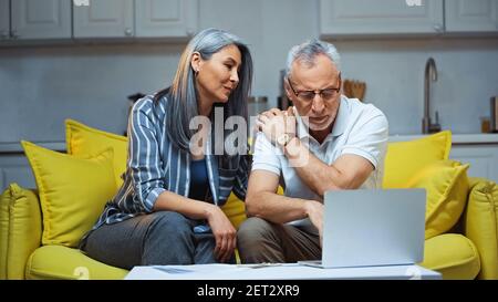 anziana coppia multietnica seduta sul divano vicino al computer portatile a casa Foto Stock