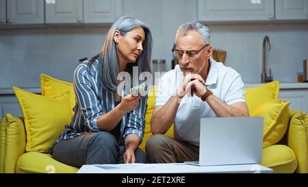 grigio capelli asiatico donna che tiene dollari vicino a computer portatile e serio marito Foto Stock