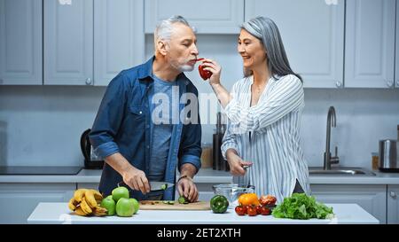 donna asiatica felice che alimenta il marito con peperone mentre si prepara colazione in cucina Foto Stock