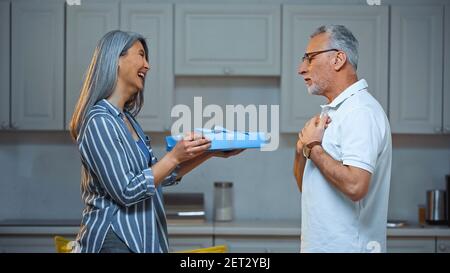 vista laterale di allegra donna asiatica che presenta dono a stupito marito anziano Foto Stock