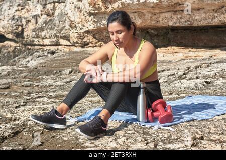 Donna latina, di mezza età, riposo, riconquista forza, mangiare, acqua potabile, dopo una sessione di ginnastica, calorie brucianti, mantenersi in forma, all'aperto vicino al mare Foto Stock