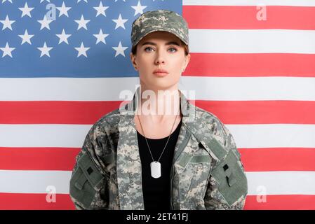 Donna in uniforme militare e cane tag guardando fotocamera vicino alla bandiera americana sullo sfondo Foto Stock