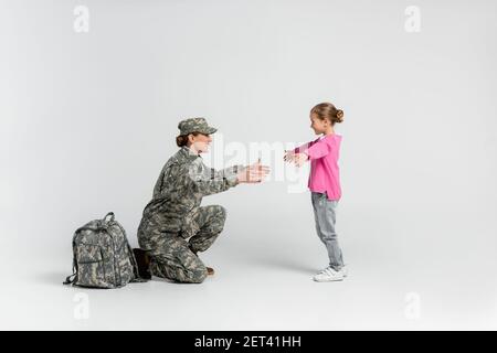 Vista laterale di madre in camuffamento inginocchiato vicino a figlia felice su sfondo grigio Foto Stock
