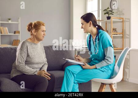 Donna matura seduta sul divano a casa che parla con l'infermiere o il medico durante la sua visita. Foto Stock