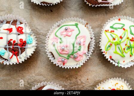 Muffin marroni al cioccolato avvolti in carta bianca e ricoperti di glassa bianca con decorazioni colorate, cotti al forno, stesi su carta da forno, a. Foto Stock