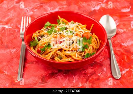 Pasta spaghetti Puttanesca in ciotola rossa con forchetta e cucchiaio su sfondo colorato. Cucina italiana. Foto Stock