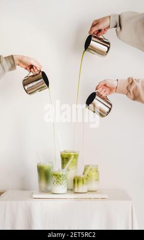 Mani femminili che versano il tè giapponese matcha ai bicchieri con latte e che fanno il latte matcha verde per la colazione a bordo su sfondo chiaro della parete. Bevande al caffè e cucina giapponese Foto Stock