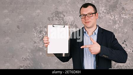 Giovane adulto sorridente uomo in una tuta e bicchieri punta il dito contro un foglio di carta vuoto contro una parete grigia di cemento. Business mockup. Banner Foto Stock