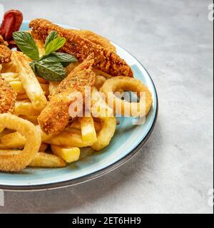 Patatine fritte, anelli di cipolla e ali di pollo in un piatto blu su sfondo blu-grigio Foto Stock