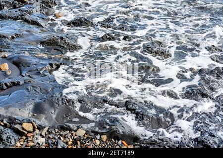 La piccola onda rotola sulla riva e spruzzi di schiuma vola in diverse direzioni. Splendido salvaschermo marino minimalista. Le onde di mare si rompono sul beac di shingle Foto Stock