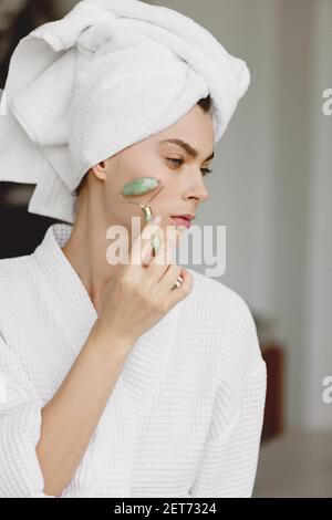 Donna attraente con asciugamano sulla testa che ottiene il massaggio del viso usando il rullo di giada. Concetto di trattamento di bellezza. Foto Stock