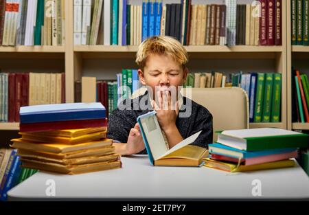 scolaro stanco che urla mentre studia, fa i compiti, seduto a tavola circondato da libri, in biblioteca Foto Stock