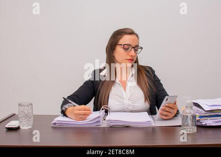Una giovane donna d'affari dai capelli marroni che indossa abiti formali e lavora in ufficio, utilizzando uno smartphone Foto Stock