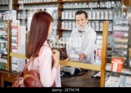 Felice sicuro farmacista maschio anziano in piedi dietro il banco e che offre la scatola della medicina, parlando con la giovane cliente della donna. Vendita di medicina Foto Stock